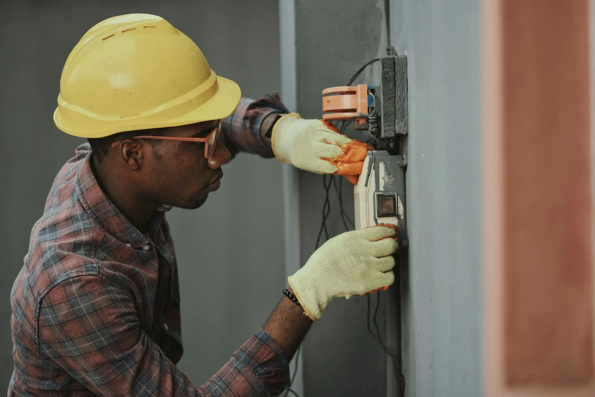 Electrician wearing a yellow hard hat checking a wall-mounted meter outdoors