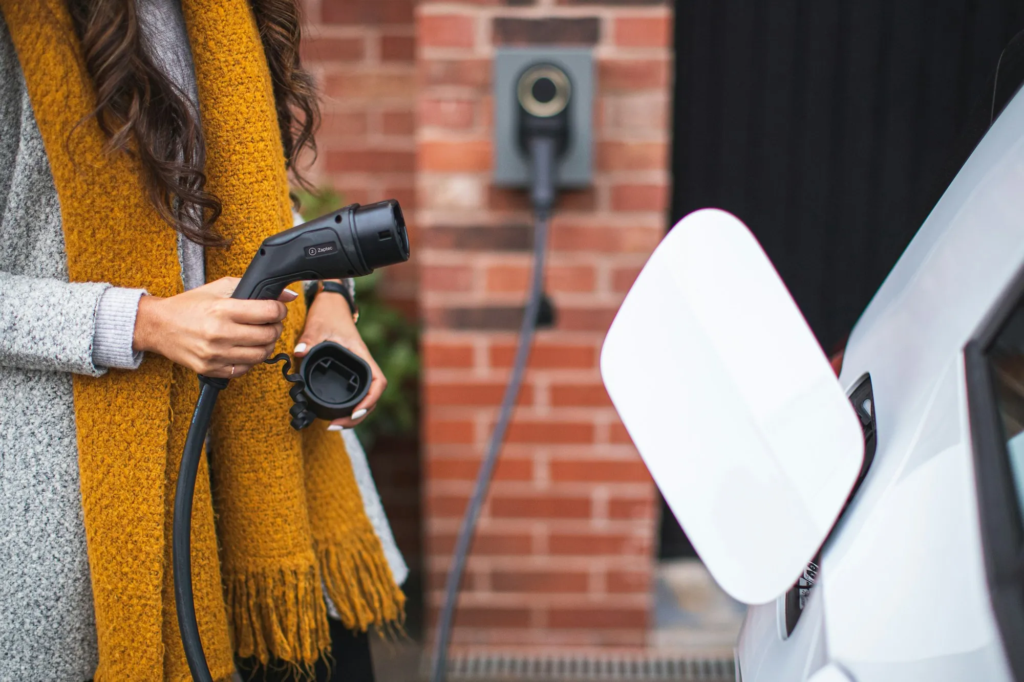 Woman in a mustard scarf holding a charger plug next to a white car